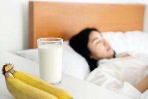 Une personne endormie dans un lit, avec une banane et un verre de lait posés en premier plan, symbolisant l’impact des glucides sur le sommeil.